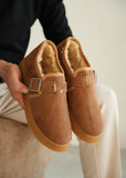 Brown suede slippers with fur lining held by a person against a neutral background